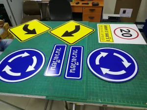 ชุดป้ายจราจรวัดพระธรรมกาย ป้ายเตือนทางโค้ง ป้ายวงเวียน ป้ายจำกัดความเร็ว 20 กม. และป้ายลดความเร็ว ถ่ายบนโต๊ะในโรงงาน