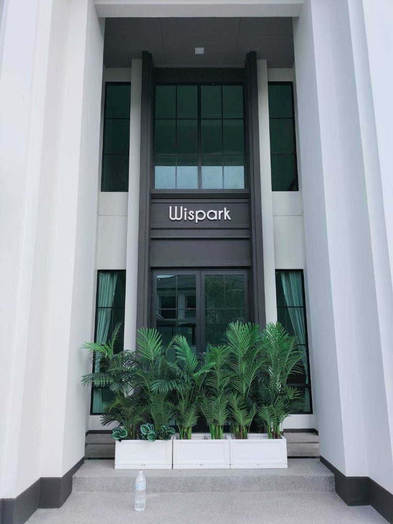 ป้ายตัวอักษรสแตนเลสนูน Wispark ติดเหนือประตูทางเข้าอาคารโมเดิร์นสีขาว เสาสูงกระจกเขียว ผลิตโดย Scinage